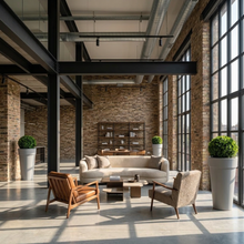 Load image into Gallery viewer, A modern industrial loft interior with exposed brick walls, large black-framed windows, and a central seating area featuring a curved light beige sofa, two accent chairs, and a low wooden coffee table. Tall light grey planters from Bigplantpots with green shrubs are placed near the windows.