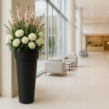Load image into Gallery viewer, A tall, slim black Bigplantpots planter placed in a modern interior setting, filled with a variety of white and pink flowers.